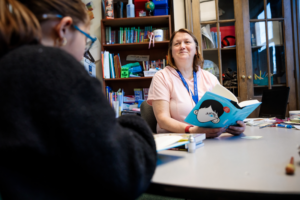 Photo of a teacher sitting at a table with a book, looking up and smiling at a student.